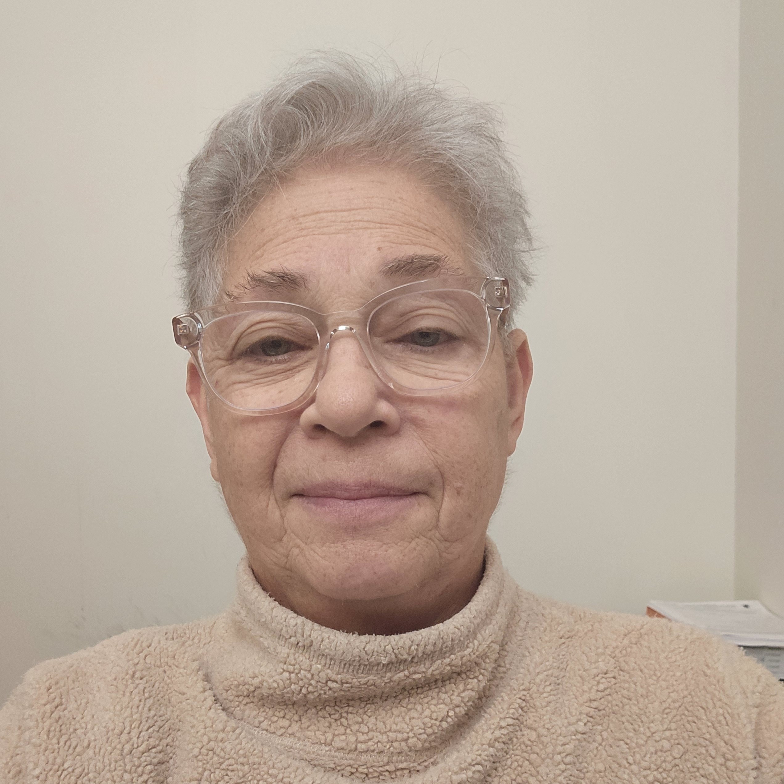 A person with short gray hair and clear-framed glasses is wearing a beige fleece turtleneck and facing the camera with a neutral expression. The background is a plain light-colored wall.